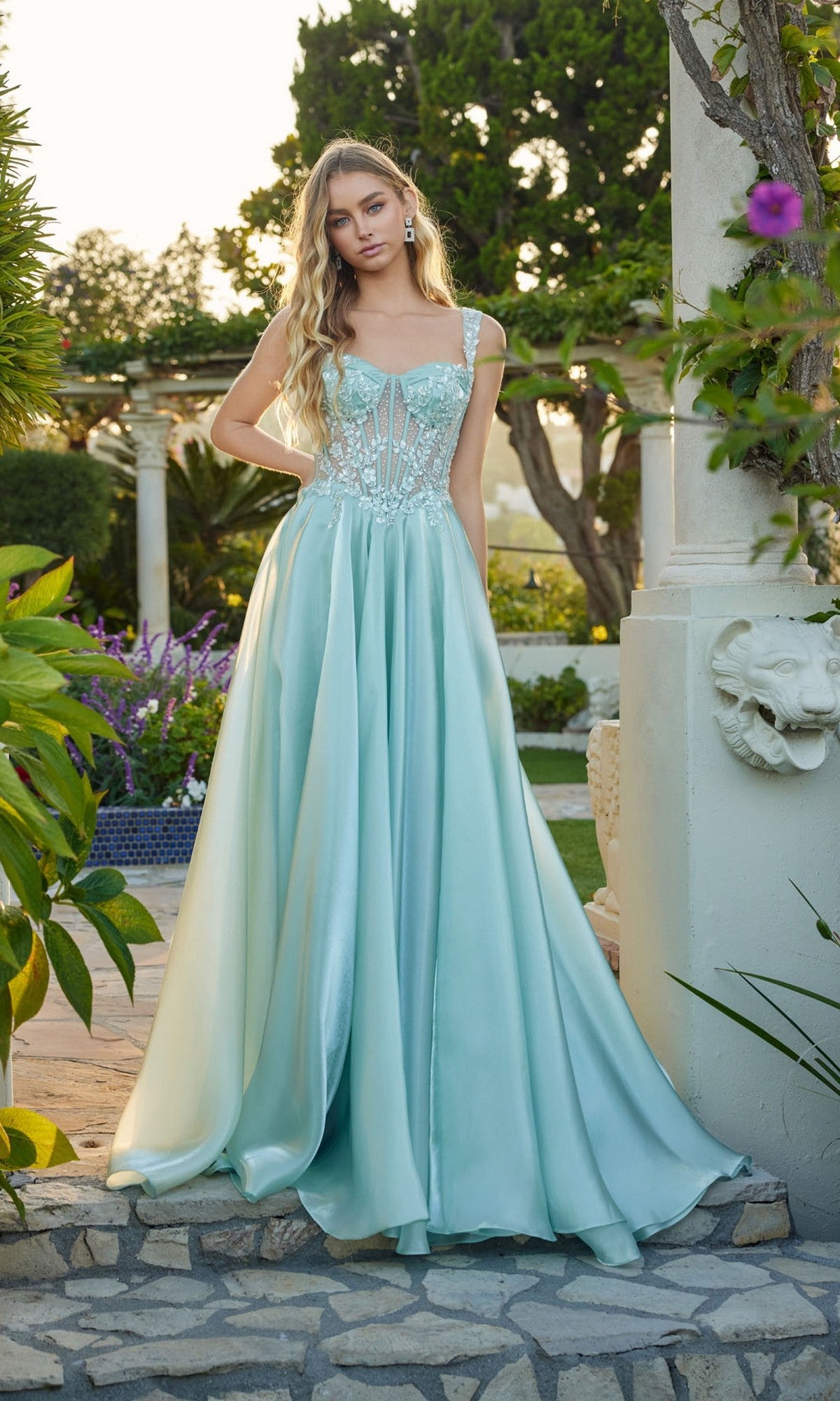 A woman wears the Juliet JT2639G, a light blue floor-length satin A-line prom dress with a sheer corset bodice, standing outdoors near greenery and stone architecture.